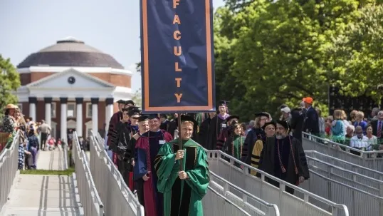 Photo of UVA faculty at final exercises wearing robes. Faculty member at the front of the line carries a banner reading FACULTY.